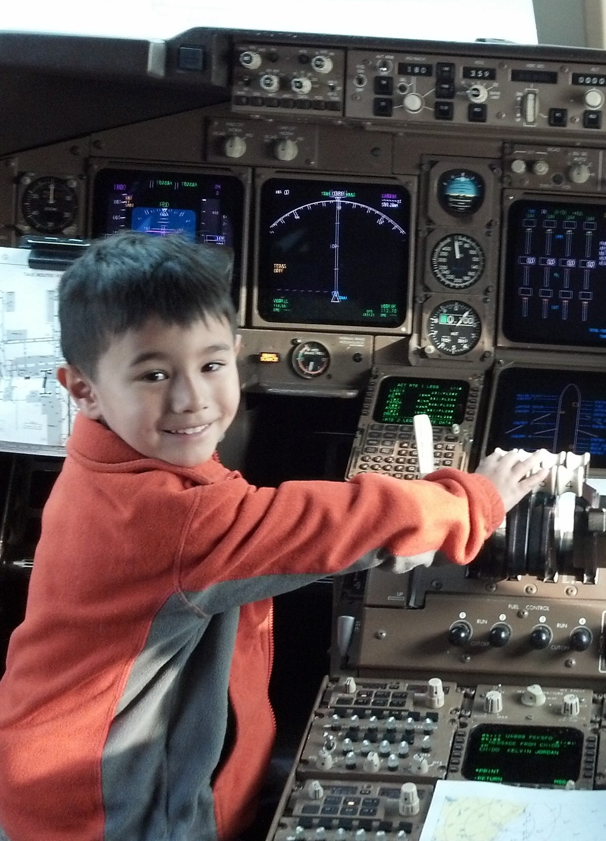 A small, Asian looking boy of about five wearing an orange fleece sits in the left-hand seat in the cockpit of a Boeing 747-400, looking back over his shoulder with a grin and his right hand on the throttle levers. A wall of instruments is visible in front of him, including multifunction displays and various knobs and dials. The kid looks as happy as can be.