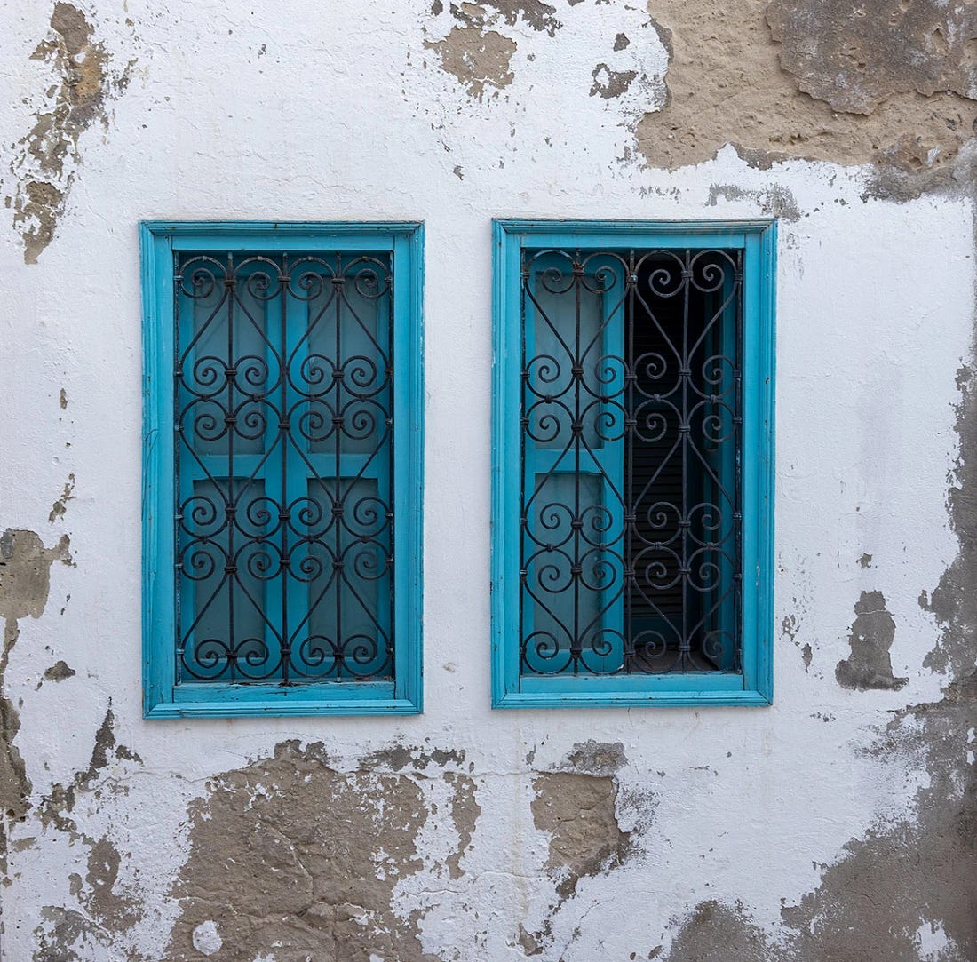 Twin turquoise-painted windows with wrought-iron grilles set into a weathered whitewashed wall in the Tunis Medina, the peeling plaster revealing layers of age and daily life.