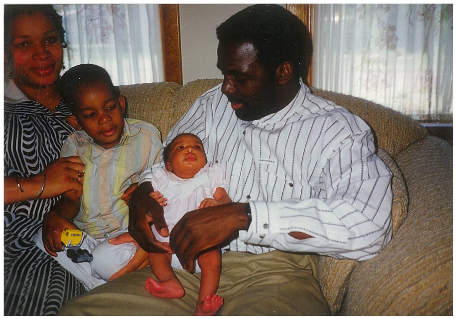 Family of 4 sitting together on the living room couch. A dark skinned African Father wearing a white pinstriped shirt and khakis sits on a couch lovingly holding a little girl infant in his arms. A a young brown-skinned boy wearing a pastel striped shirt sits to his right, gazing curiously at the infant. A light-skinned Afrolatina woman wearing a black and white striped dress gazes into the camera while holding the arm of the boy.