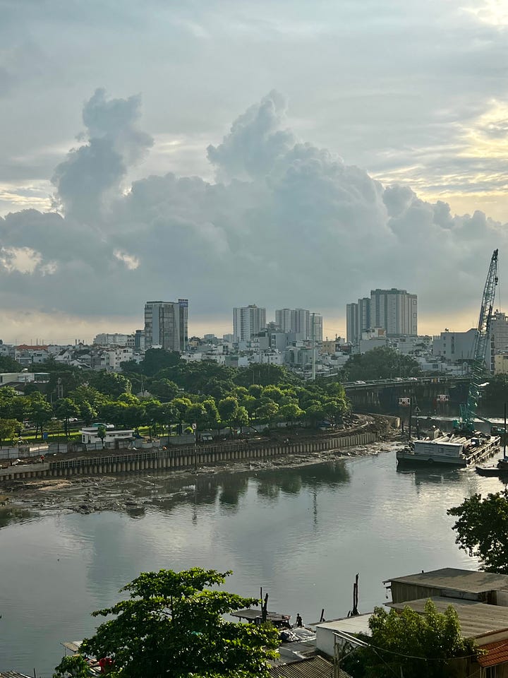 Daytime views of Ho Chi Minh City: a riverside skyline with high-rise buildings under dramatic clouds and construction along the riverbank, and a street scene with motorbikes passing a small “Com Tam Bi Bo” eatery while people dine on red plastic chairs and a dog sits near the curb.