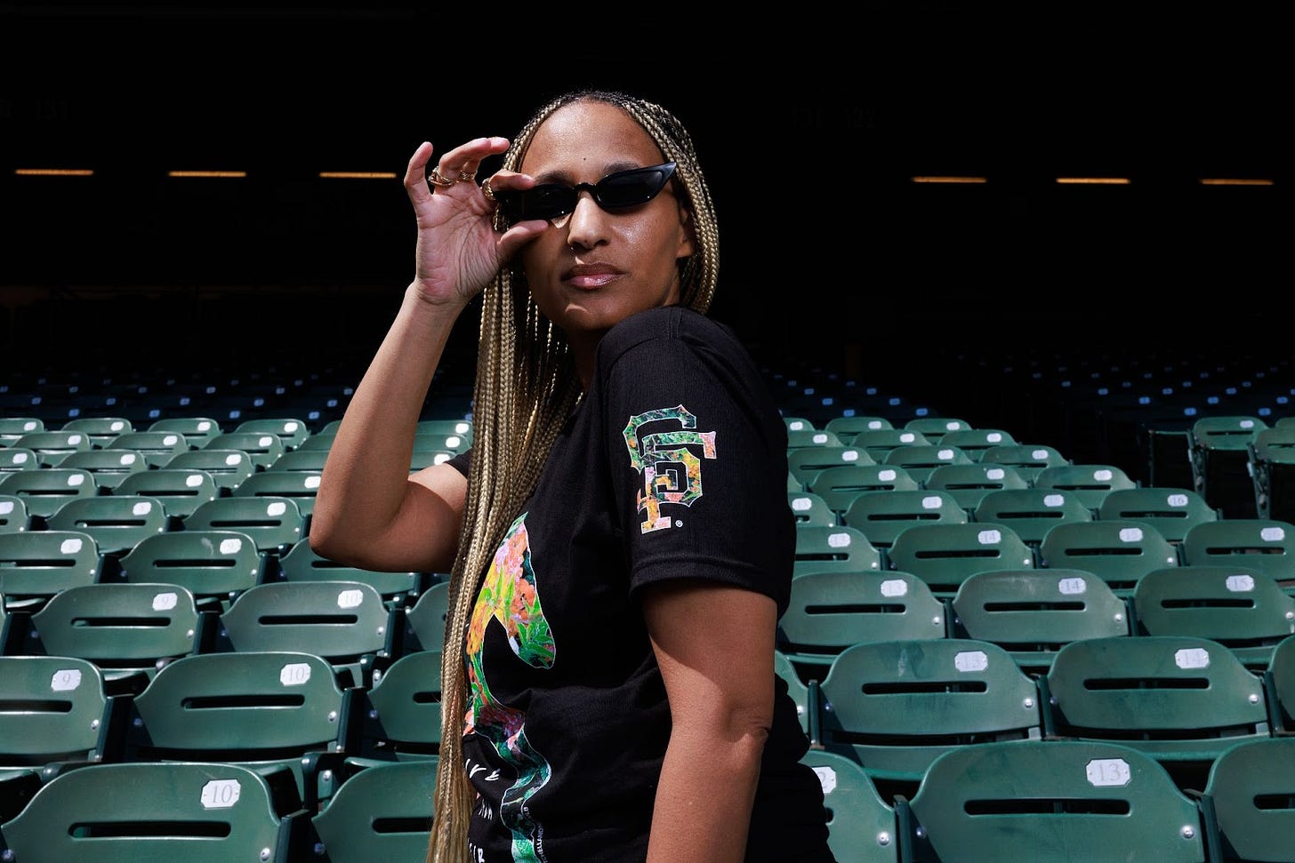 Standing in a sea of green ballpark seats Dr. Cadet wears her Flowers black t-shirt showing the SF floral logo on the side of the sleeve. Looking in the direction of the branded shoulder Dr. Cadet has one hand of her black cat eye sunglasses giving you the “yes I know I’m the shit” pose with her golden braids tucked between her arm and award winning cleavage