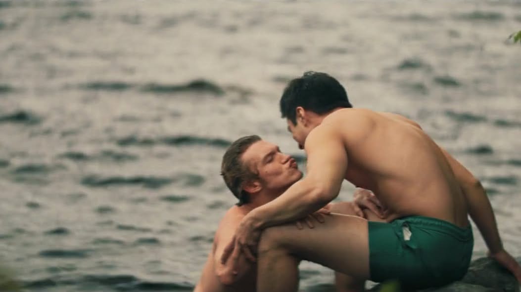 a still from Heated Rivalry, episode 6: Shane, an Asian Canadian man sits on a rock in the water in a green bathing suit; Ilya, a white man, leans toward Shane, lips puckered, asking for a kiss