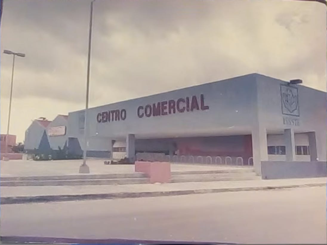 A faded color photo of a low white concrete building with CENTRO COMERCIAL in red letters across the front and the ISSSTE crest on the wall, an empty parking lot, and an overcast sky.