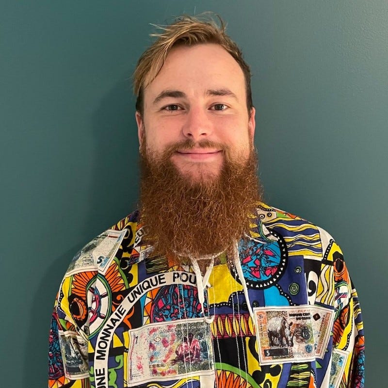 This is a portrait of Adrian Wood. He has a full, majestic beard and is smiling slightly. He is wearing a colorful, patterned shirt with various graphic designs and text. The background is a solid, muted green color.