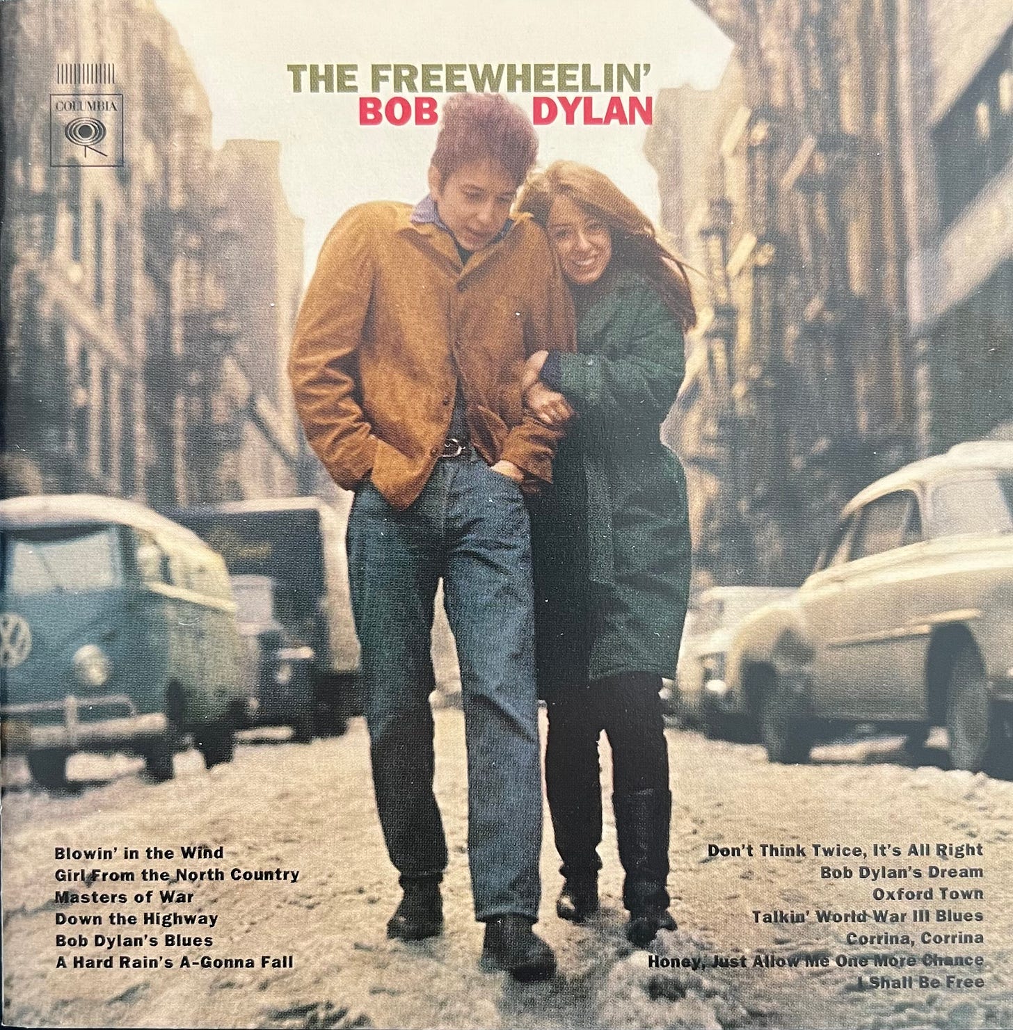 Image depicts the cover of Bob Dylan's album, The Freewheelin Bob Dylan. The image shows Bob Dylan walking arm in arm with his then girlfriend Suze Rotolo in the middle of a New York street in the early 1960s. There are cars on both sides of the street.