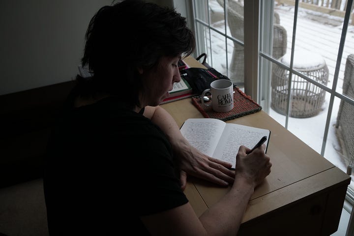 the author, a masculine white person with short brunette hair, stands outside by a tree; snow blankets the background. next, the camera peers over their shoulder as they write in a notebook at a desk that oversees the wintery landscape