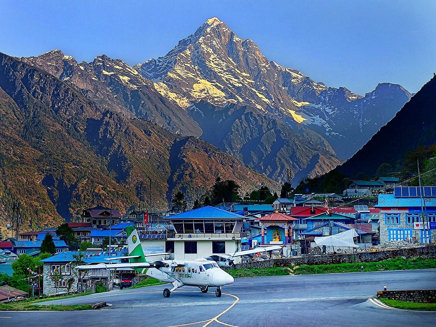 Foreground shows a dual-engine prop plane on the taxiway at Lukla airport, with the village visible behind. Spanning across the background is a snow covered mountain peak bathed in sunshine.