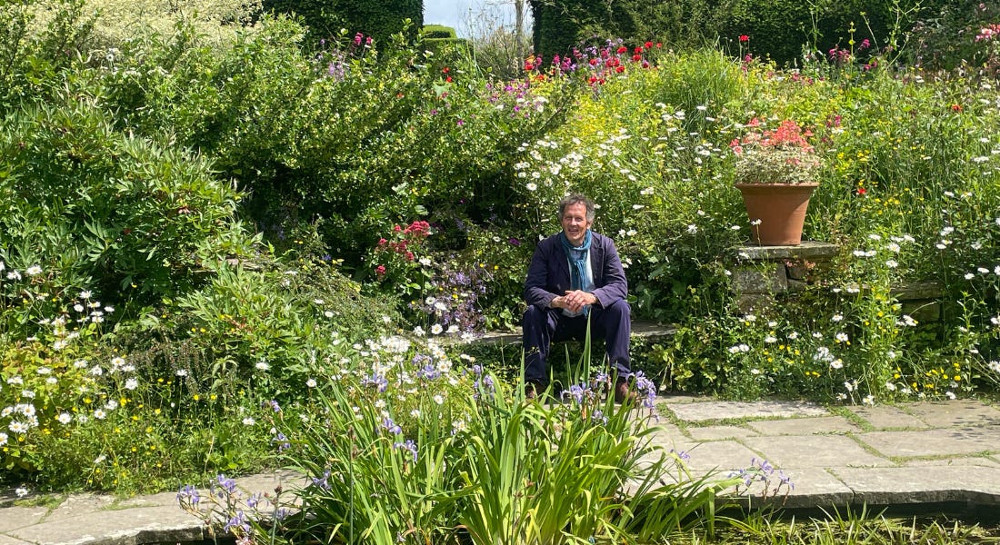 Monty Don wearing a blue outfit sat in a garden with bright green bushes and white, pink, and purple flowers around him Monty Don wearing a blue outfit sat in a garden with bright green bushes and white, pink, and purple flowers around him