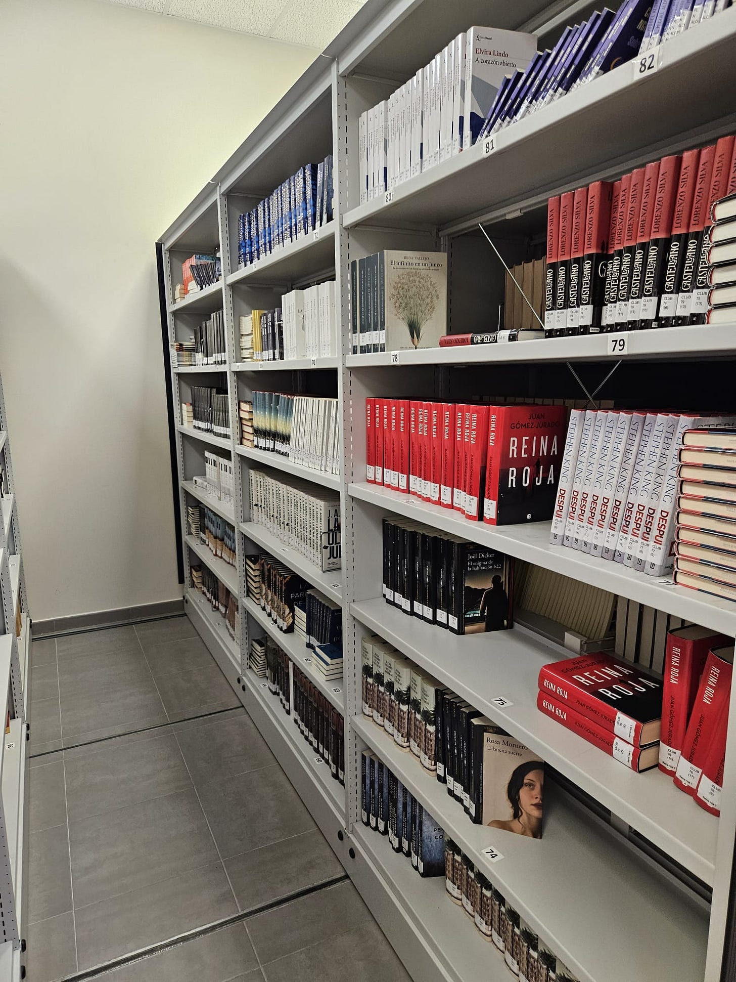 A imagem mostra uma seção de estantes de livros em uma biblioteca, organizada com prateleiras metálicas de cor clara, onde estão dispostos vários livros. As prateleiras estão alinhadas de forma ordenada, com etiquetas visíveis na parte inferior de cada prateleira, indicando a classificação dos livros, como num sistema de catalogação.  Os livros são predominantemente de cores vermelha, branca e preta, criando um contraste interessante. Na estante à frente, há livros com capas vermelhas e títulos visíveis, como Reina Roja de Juan Gómez-Jurado. Entre os livros vermelhos, há também algumas edições em cores mais neutras, como o branco e o preto, que estão organizados de forma regular. No meio de uma das prateleiras, há uma capa de livro destacada, com uma imagem de uma mulher na capa, possivelmente parte de uma obra literária.  A iluminação suave ilumina as prateleiras, e o piso é de azulejos cinza, dando uma sensação de um ambiente moderno e bem organizado. No fundo, é possível ver a continuidade das estantes, criando uma sensação de profundidade e espaço. A cena transmite uma atmosfera calma e propícia à leitura.