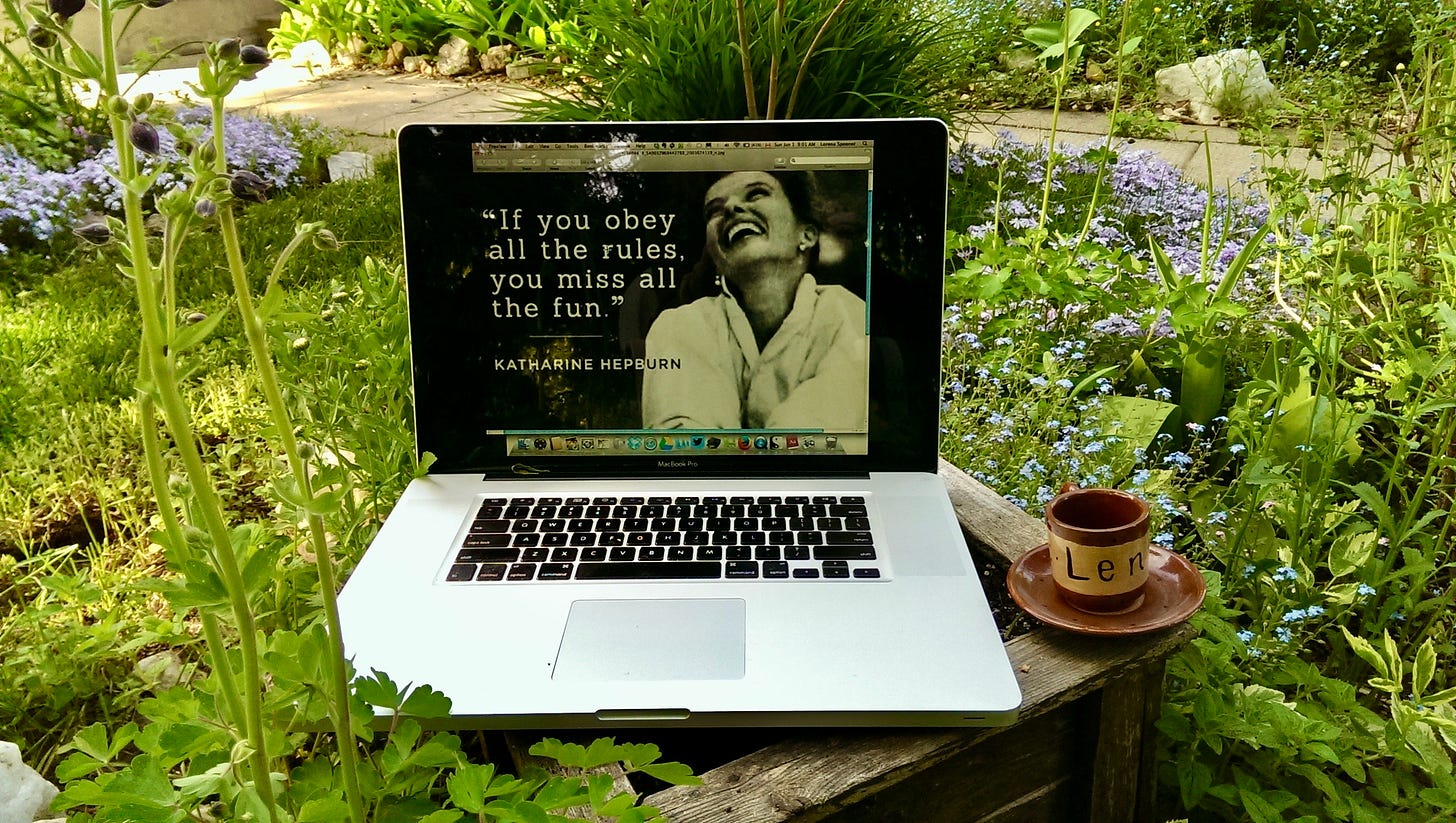 A laptop on a small garden table shows a Katharine Hepburn quote on the screen, beside a clay cup labeled “Leni,” surrounded by flowers and greenery.