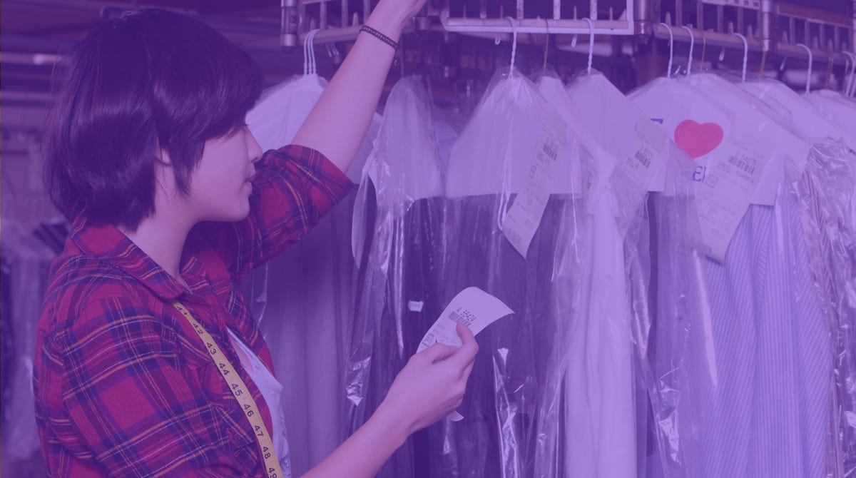 A person with short dark hair and a red plaid shirt checks a customer receipt to find their dry cleaning. They stand next to a rack of clothes that have been dry cleaned.