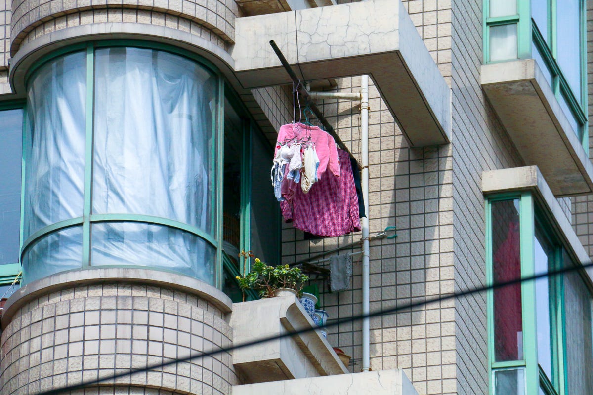 A cluster of garments, mostly pinks and whites, hang from a cantilevered metal pole on a tiled high-rise corner. A quiet domestic act defying gray skies.
