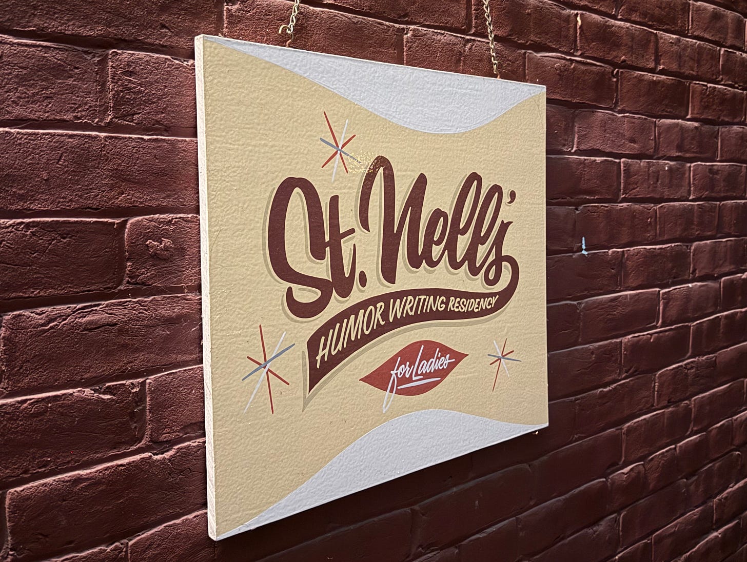 Sign hanging against a brick wall that says "St. Nell's Humor Writing Residency For Ladies"