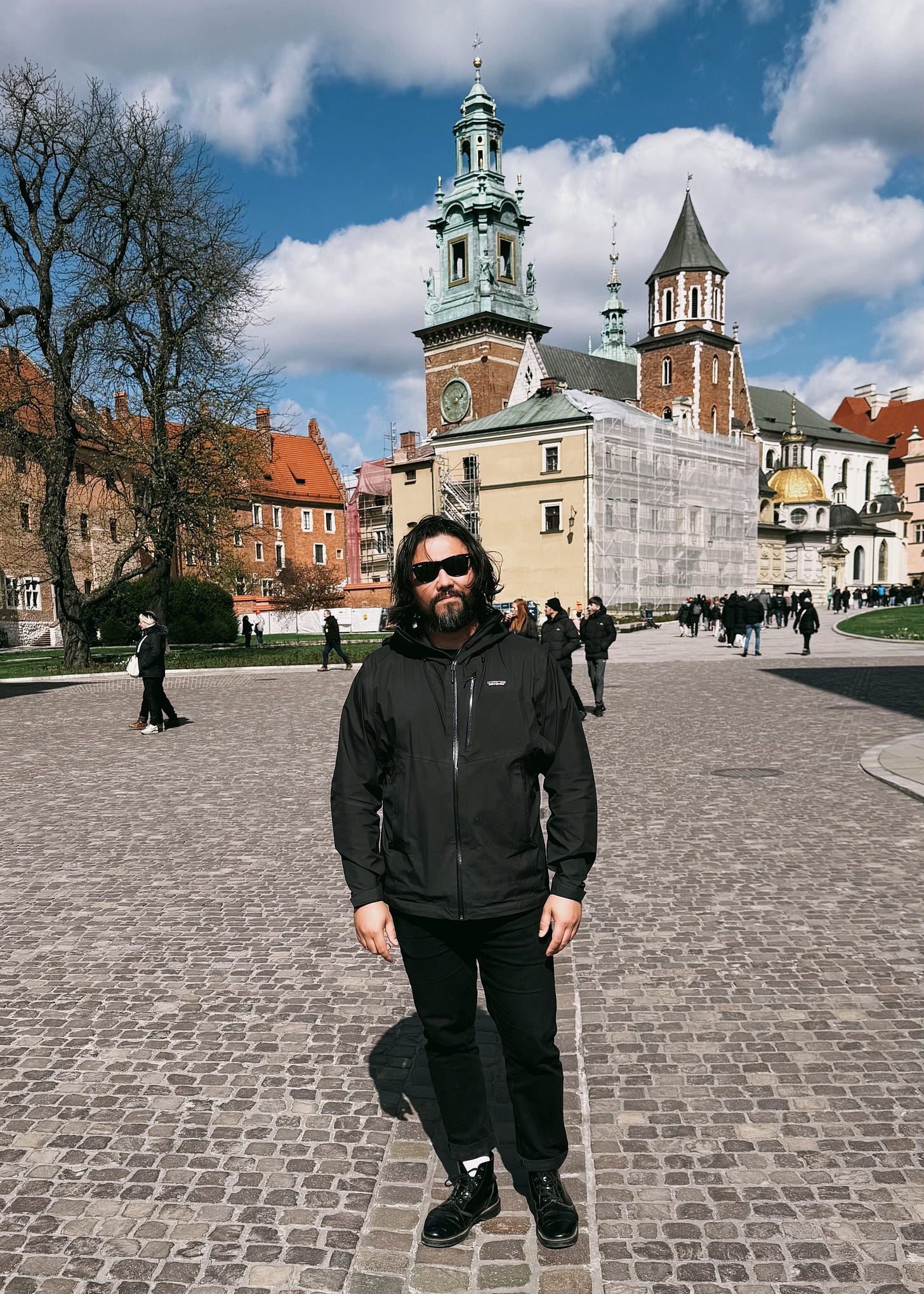 A mixed race man with long black hair dressed in black stands in front of a medieval catherdral