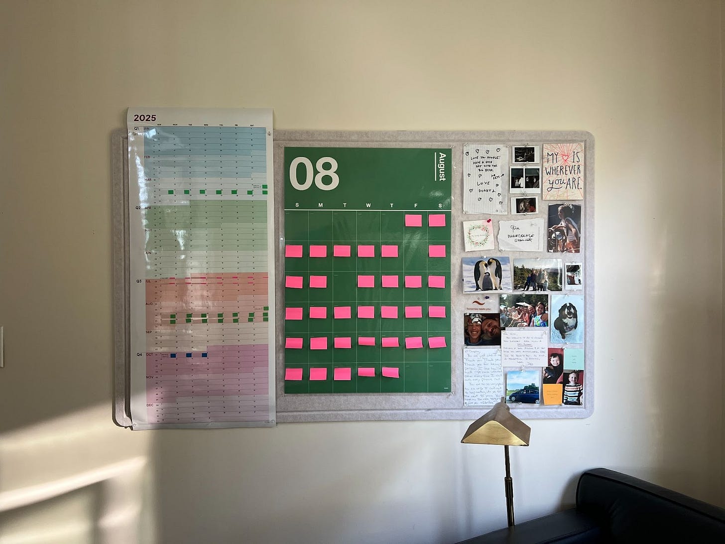 A photograph of a calendar wall with a big green monthly calendar covered in pink sticky notes, next to a board of inspirational cards and photos of Kate, her husband Dave and her dogs, Waffle and Tugboat.