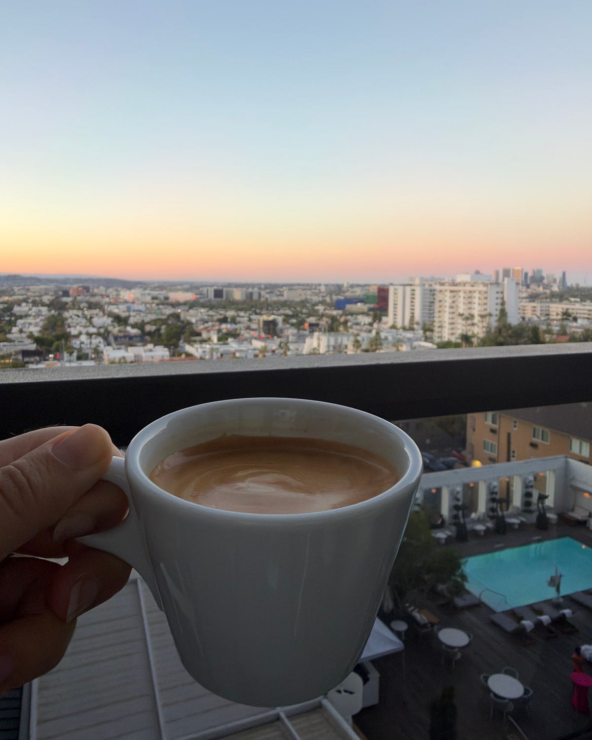 A cup of coffee held up against the Los Angeles skyline at sunset, with a hotel pool far below the balcony A cup of coffee held up against the Los Angeles skyline at sunset, with a hotel pool far below the balcony