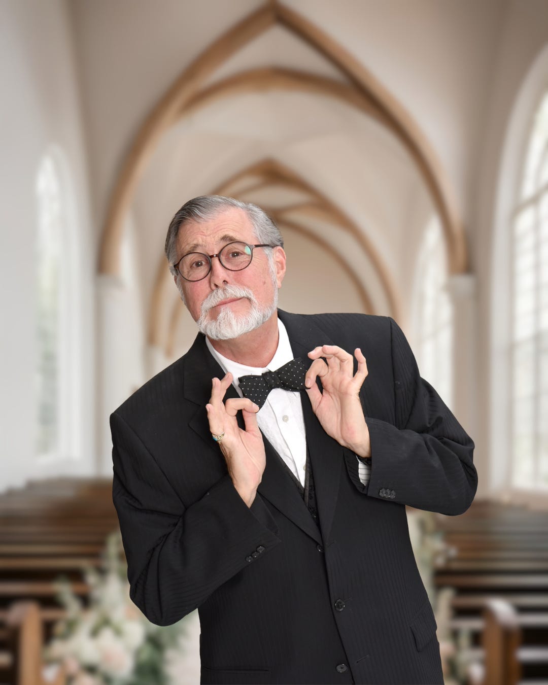A distinguished, bespectacled man with a neatly trimmed grey beard and mustache is featured from the waist up, dressed in a classic black tuxedo with a crisp white shirt. His hands are positioned near his black polka-dotted bow tie, as if he's adjusting it or drawing attention to it. He wears dark-rimmed glasses and gazes directly at the viewer with a slightly amused or inquisitive expression. The background is a softly blurred interior of a church aisle, with arched ceilings and rows of wooden pews visible on either side, suggesting a formal or celebratory occasion.
