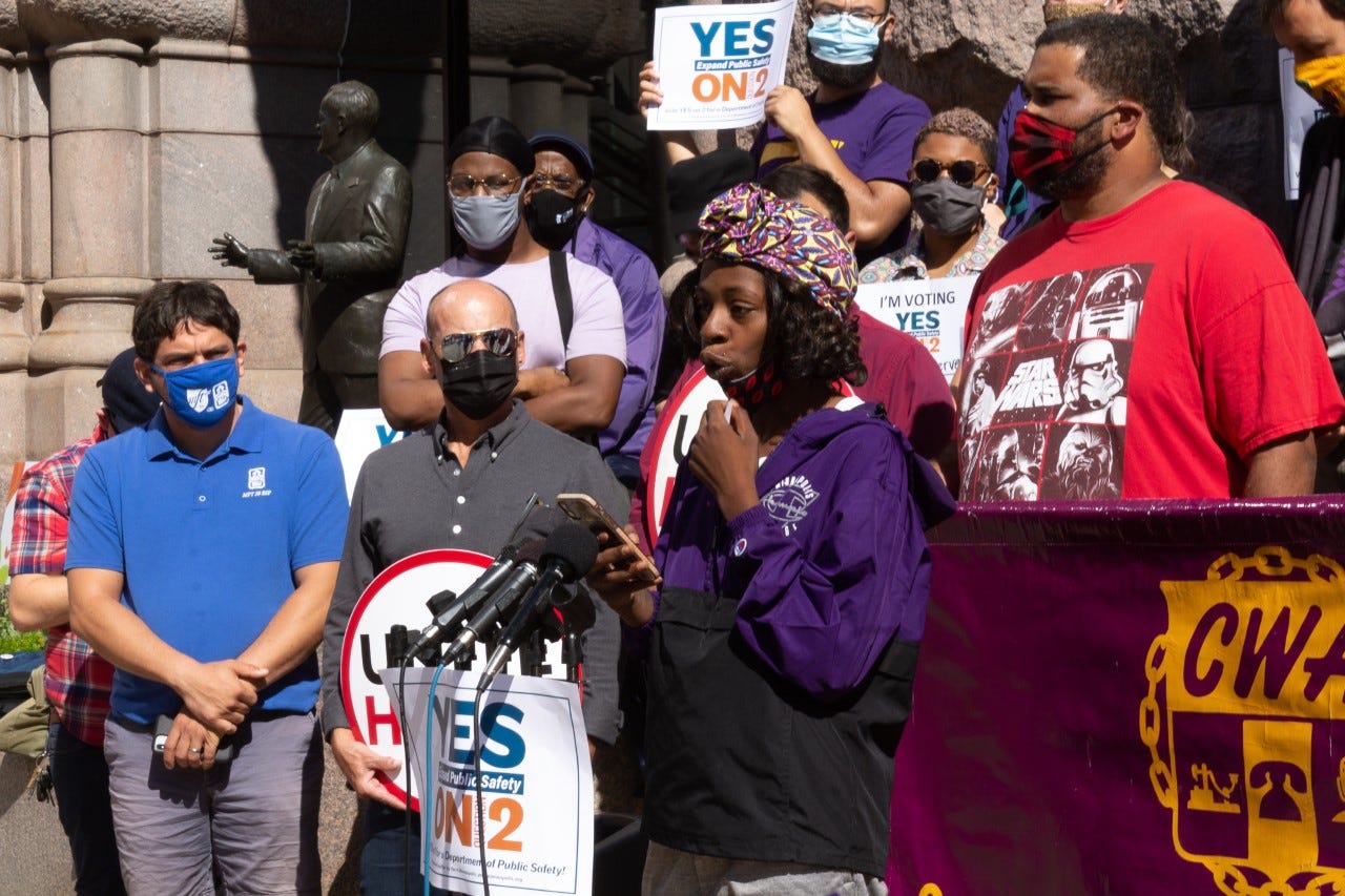 Lexi Collins, a Black woman wearing a purple and black windbreaker and purple and yellow headscarf addresses a crowd in front of a group of people Lexi Collins, a Black woman wearing a purple and black windbreaker and purple and yellow headscarf addresses a crowd in front of a group of people