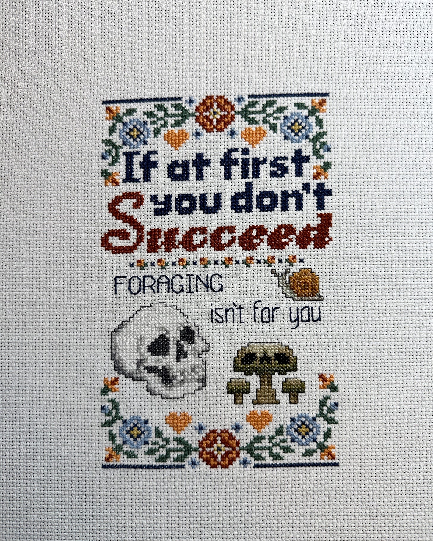 Cross stitch sampler with text that says "If at first you don't succeed, Foraging isn't for you." There is a picture of a skull and a poisonous mushroom at the bottom of the sampler.