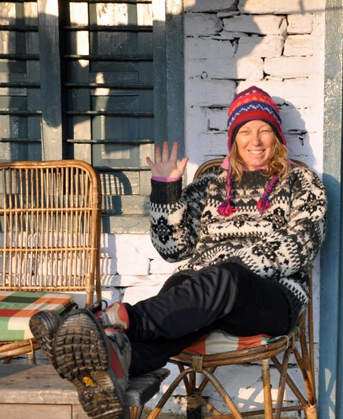 White woman with blond hair is wearing colorful yak-knit hat, sweater, and socks. She is sitting in front of a wooden shack. 