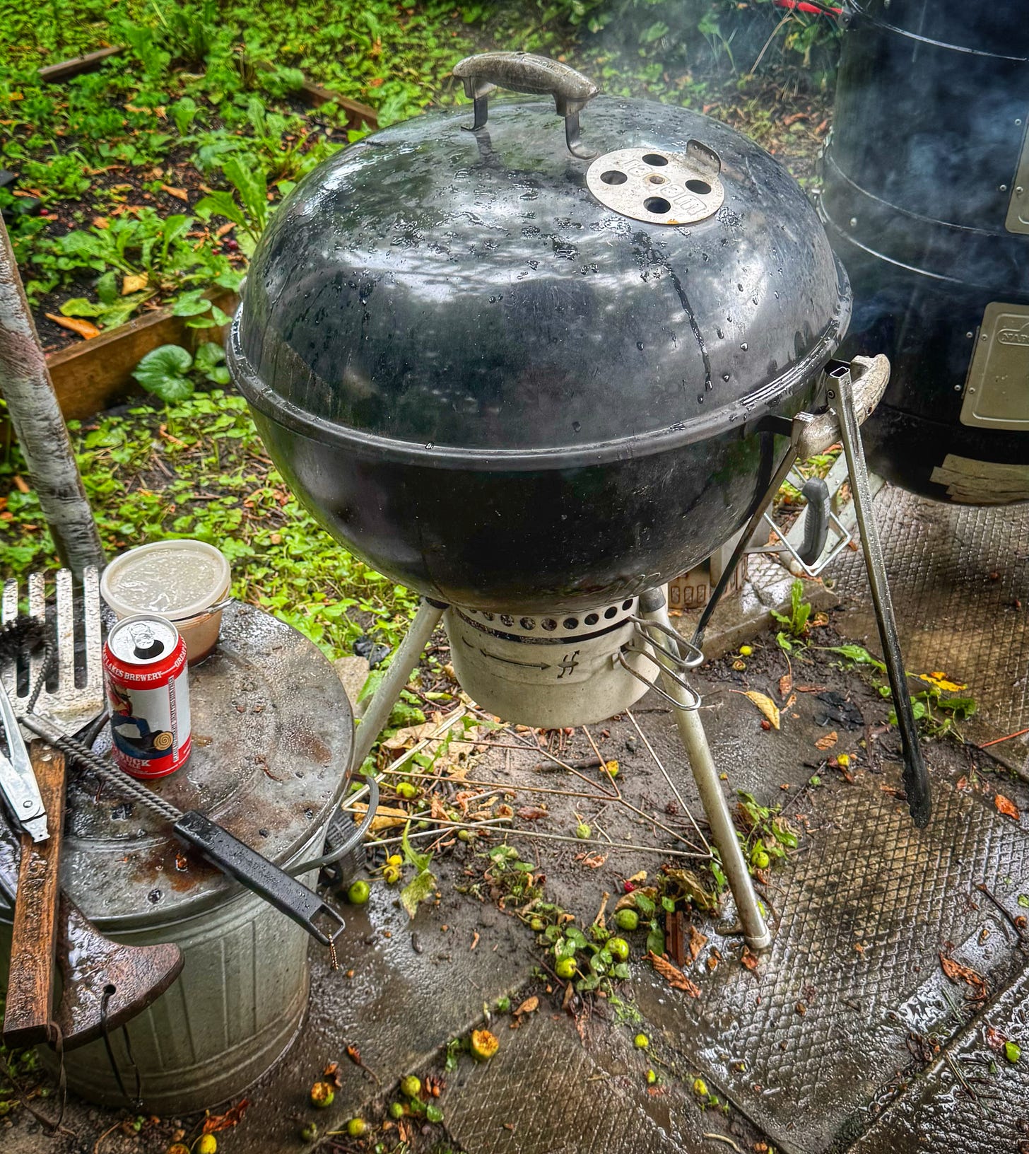 A Weber kettle grill with the lid on. Smoke is emerging from the vent holes. Around the grill, the ground and other areas look wet, but it is not clear that it is raining as heavily as it actually was. 