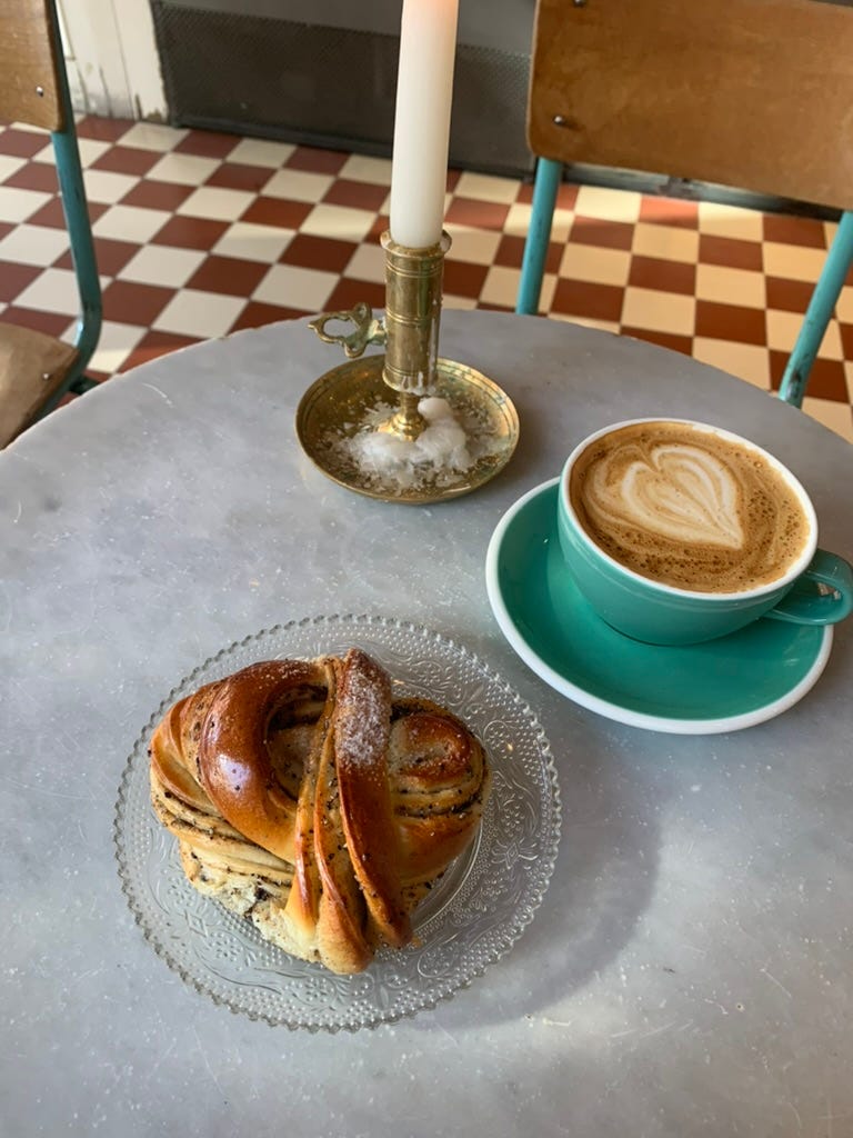 Three-quarter angle of a circular white marble table with a knotted cinnamon pastry on an ornate glass plate next to a frothy cup of coffee in a mint green cup and saucer, near a tall white candle stick. In the background the cafe had a dark red and cream checked tile floor.