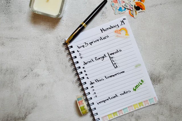 An open spiral notebook on a textured surface showing a handwritten daily planner for Monday the 24th, with sections for top three priorities, meals, and important notes, alongside a candle and colorful stickers, representing the standard time management approach that creative capacity mapping asks artists to reconsider.