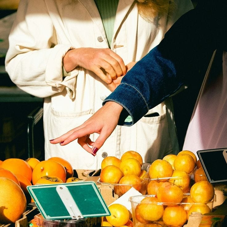 This may contain: two people reaching for oranges at an outdoor market table with other fruit on display