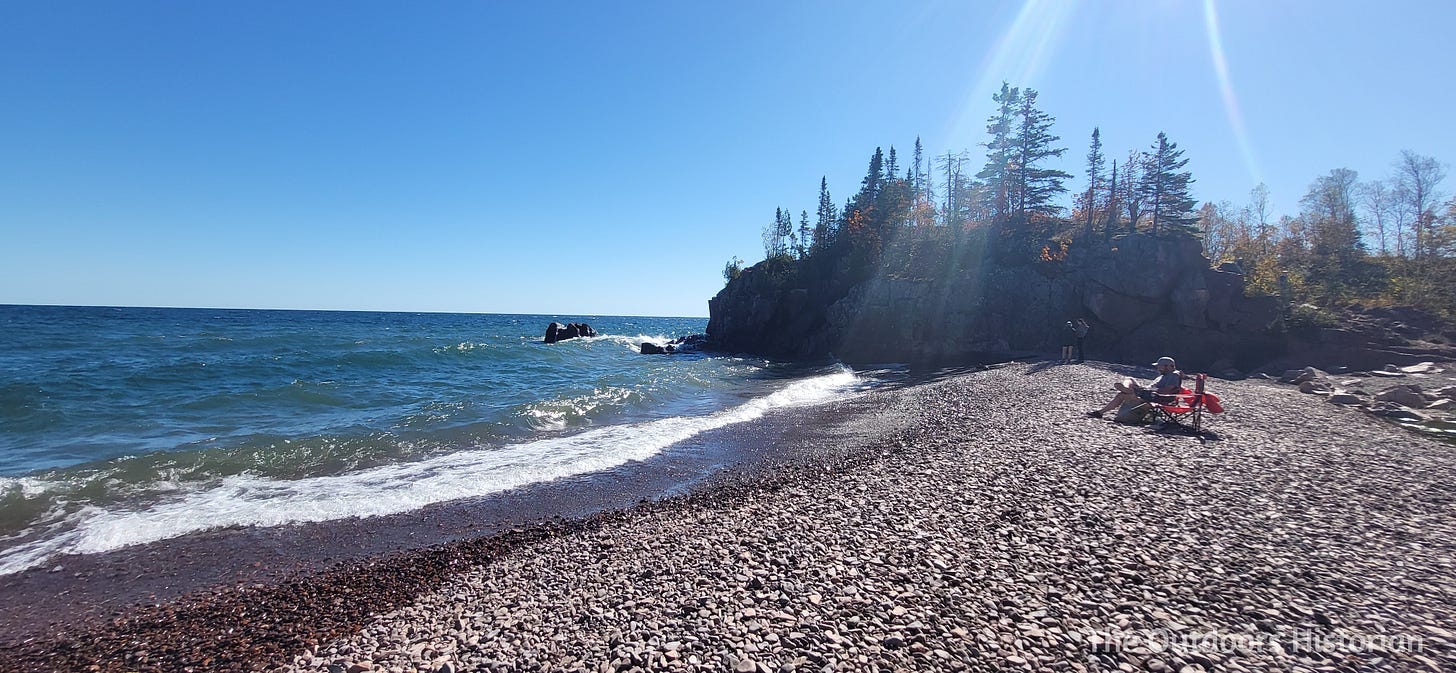 A person sits on a chair on a beach made of black stone and rock on a sunny day.