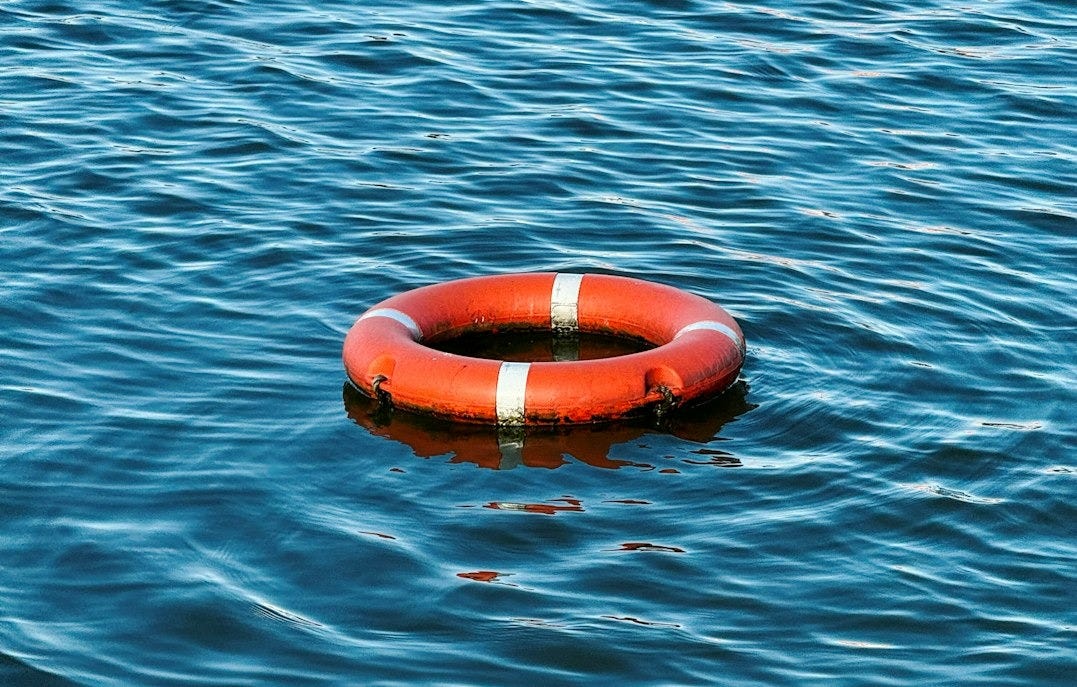 An orange lifebuoy floats on rippling blue water. (there's a buildup of algae on the bottom so it may be anchored in place?) An orange lifebuoy floats on rippling blue water. (there's a buildup of algae on the bottom so it may be anchored in place?)