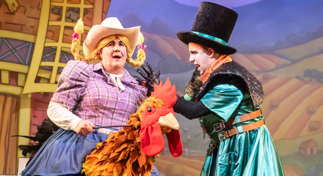 Two male performers on stage - one wearing a cowboy hat, pigtails, a checked shirt and skirt riding a chicken and one wearing a black top hat and green silky coat
