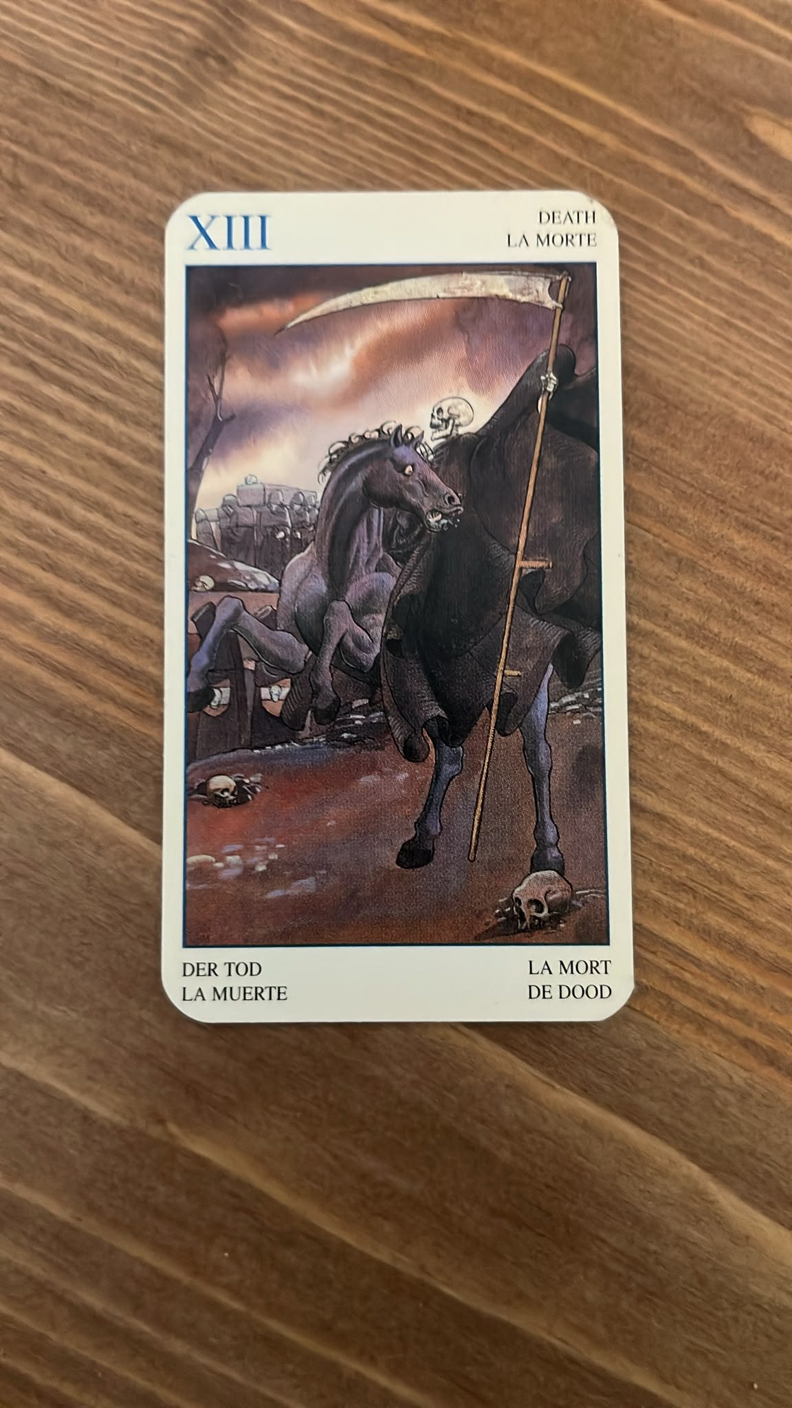 The Death tarot card lies on a wooden table, depicting a skeleton carrying a scythe while riding a dark horse through a barren landscape. The Death tarot card lies on a wooden table, depicting a skeleton carrying a scythe while riding a dark horse through a barren landscape.