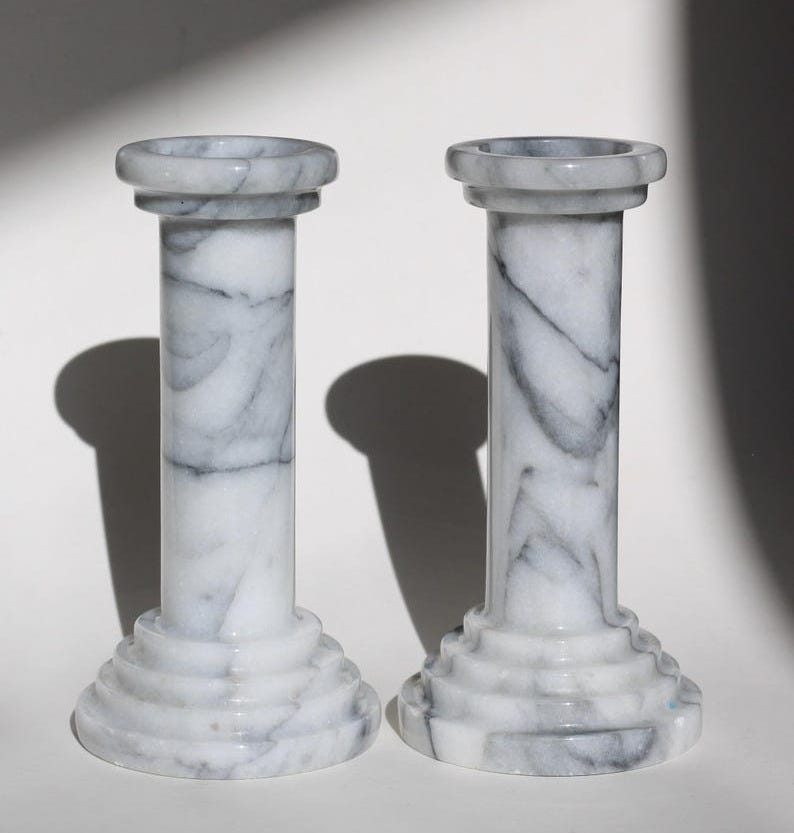 May include: Two white marble candle holders with a gray marbling pattern. The candle holders are shaped like columns with a wide base and a narrow top. The candle holders are on a white surface.