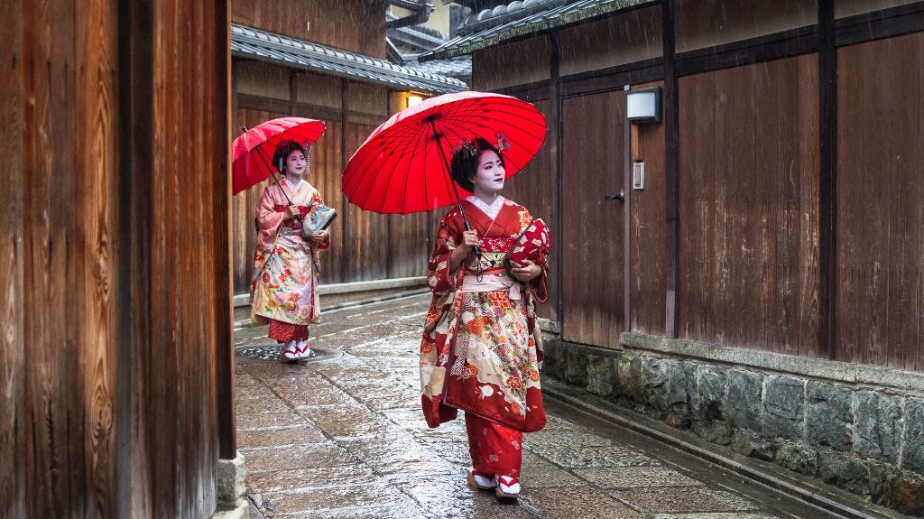 Two women dressed as geishas walk down a narrow, rain-soaked alley in Japan, each holding a bright red traditional umbrella. They wear ornate kimonos with floral patterns and wooden sandals, and the wooden buildings around them add to the historical atmosphere.