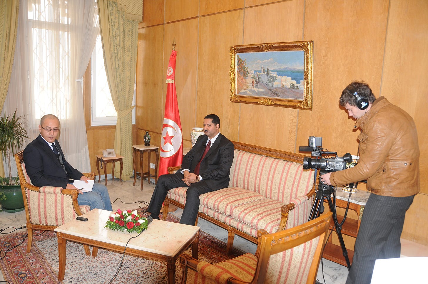 Two men sit in armchairs in a formal reception room beside a Tunisian flag while a cameraman films the conversation. Two men sit in armchairs in a formal reception room beside a Tunisian flag while a cameraman films the conversation.