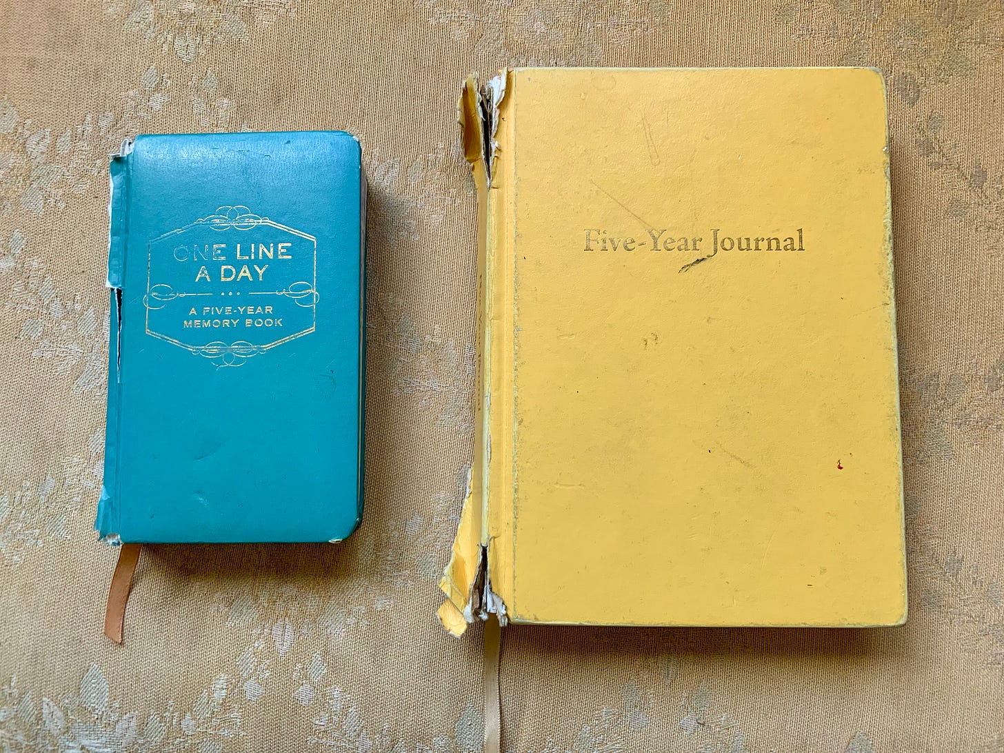 A photo taken from above of two journals against faded orange floral upholstery. On the left, a smaller blue one with the words “One Line A Day: A Five-Year Memory Book” printed on the cover. On the right, a larger yellow journal with the words “Five-Year-Journal” printed on the cover.