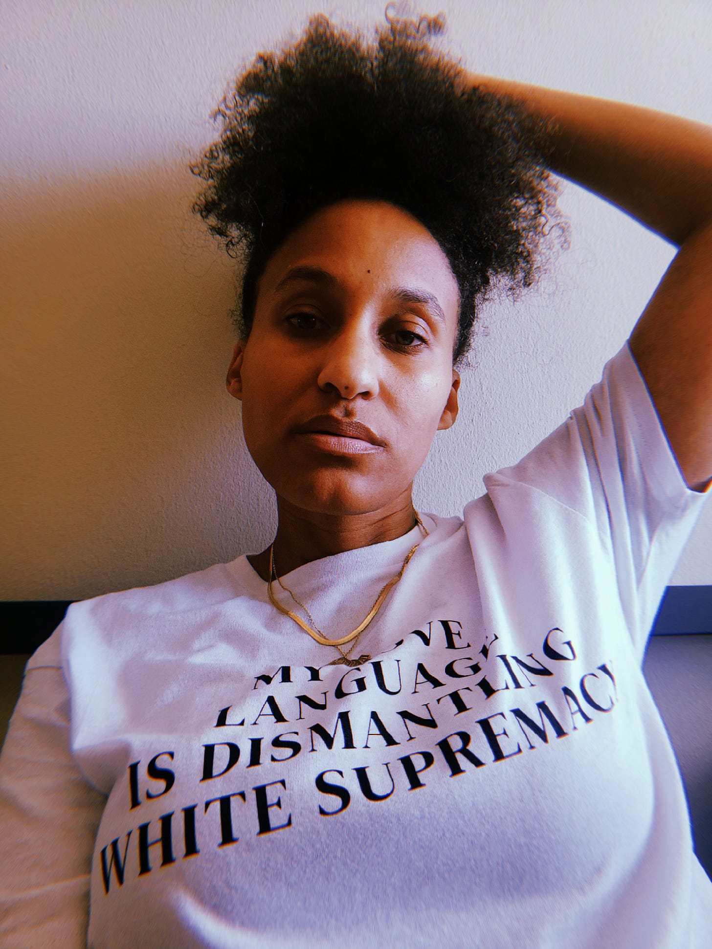 Dr. Cadet captured herself seated wearing her own shirt that says My Love Language Is Dismantling White Supremacy, which is white of course. With one hand on her head she looks directly at you as the exhauted Black disabled woman that she is.