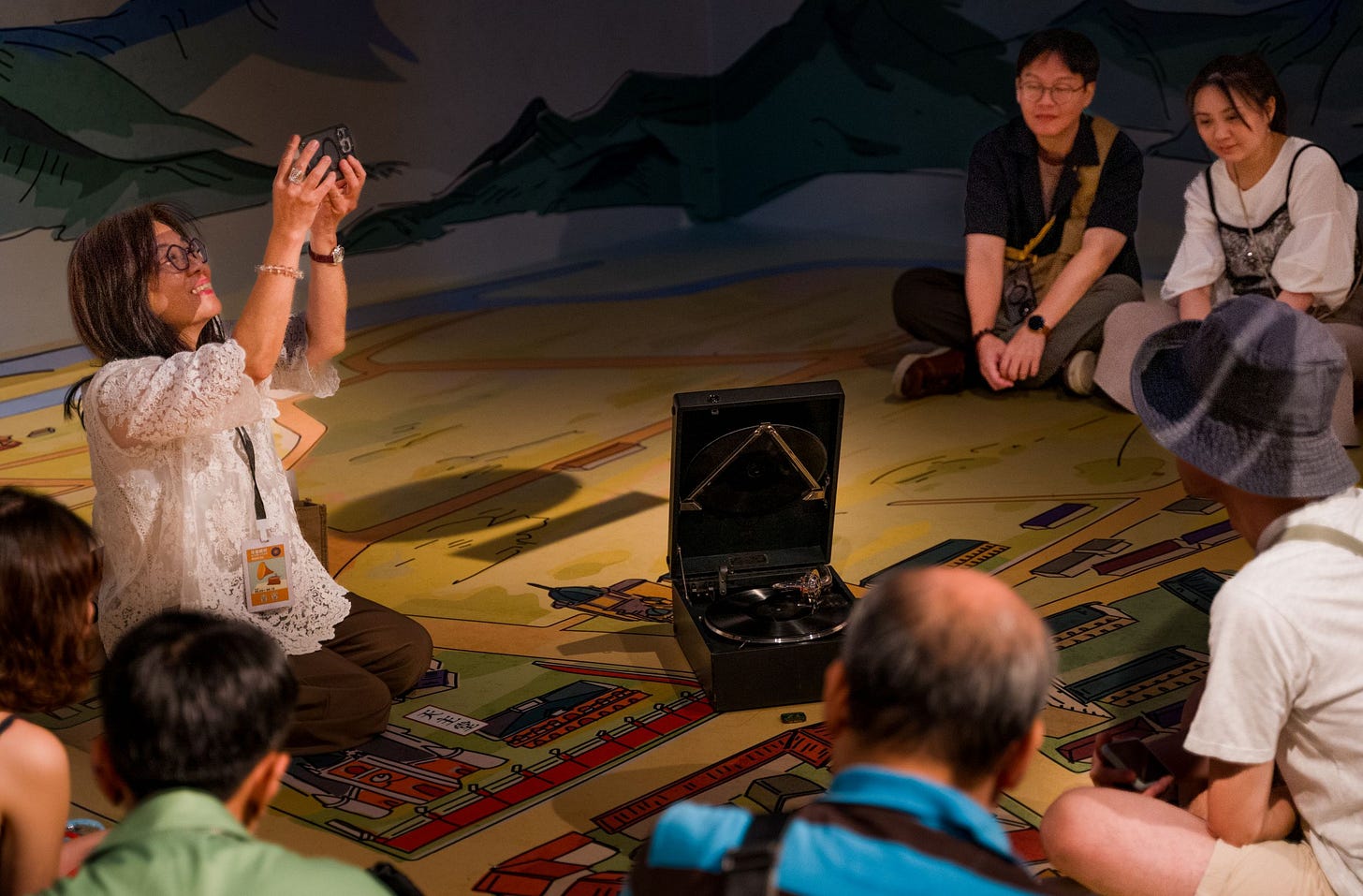 An elegantly dressed woman sitting on a colorfully illustrated floor takes photos of a group of people clustered around a portable gramophone player