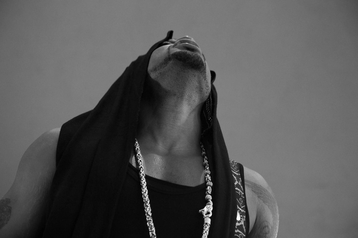 A black and white photograph of D'Angelo wearing a black sleeveless shirt with a hood draped over his head and a large necklace hanging from his neck. He's looking upwards so his face is partly obscured. The background is plain and uncluttered, emphasizing the dramatic pose. A black and white photograph of D'Angelo wearing a black sleeveless shirt with a hood draped over his head and a large necklace hanging from his neck. He's looking upwards so his face is partly obscured. The background is plain and uncluttered, emphasizing the dramatic pose.