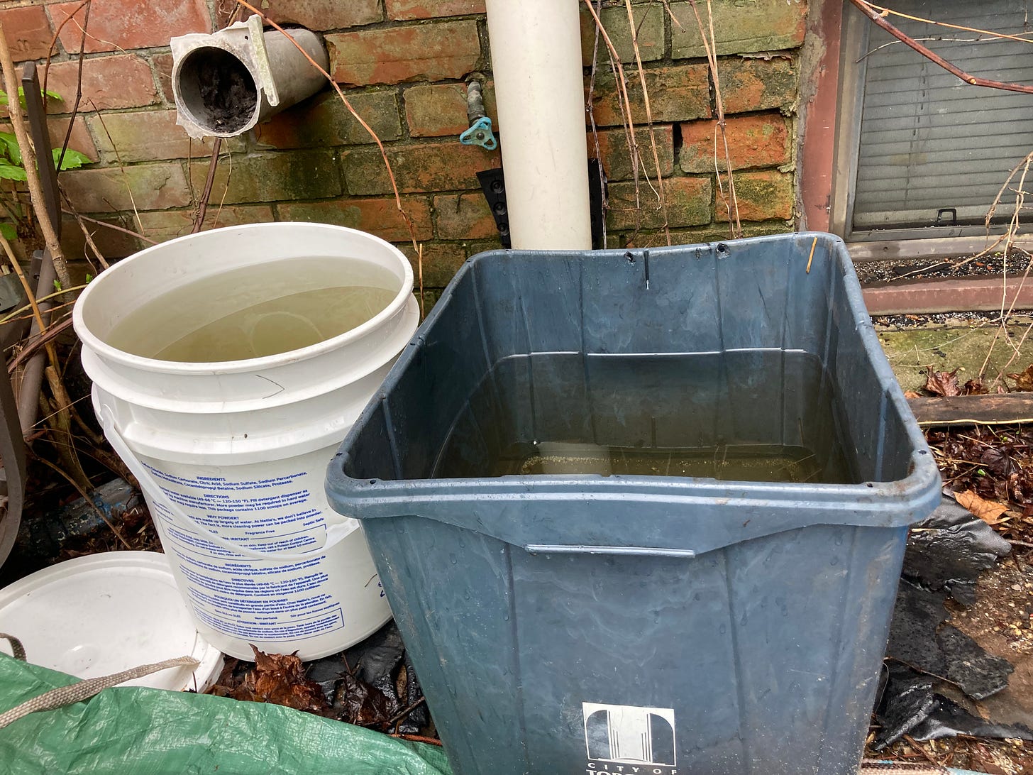 In front of a brick wall sit a large bucket and a square recycling bin, each filled with rainwater. In front of a brick wall sit a large bucket and a square recycling bin, each filled with rainwater.