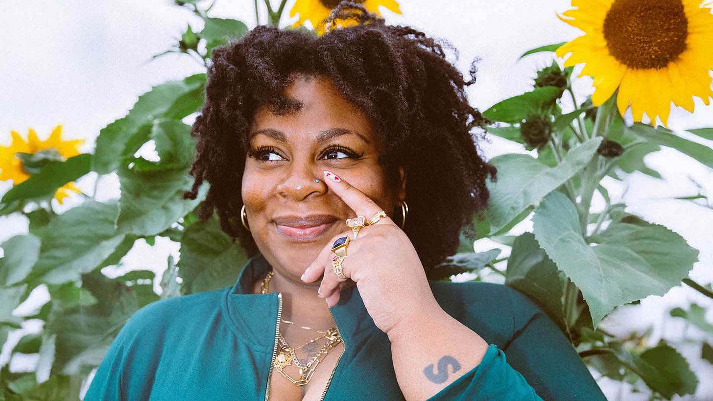 An image of Candice Carty-Williams, a Black British woman with gold jewellery and a blue-green top. She is smiling and looking off to the side, touching her face with one hand. A tattoo of an S is visible on her wrist.