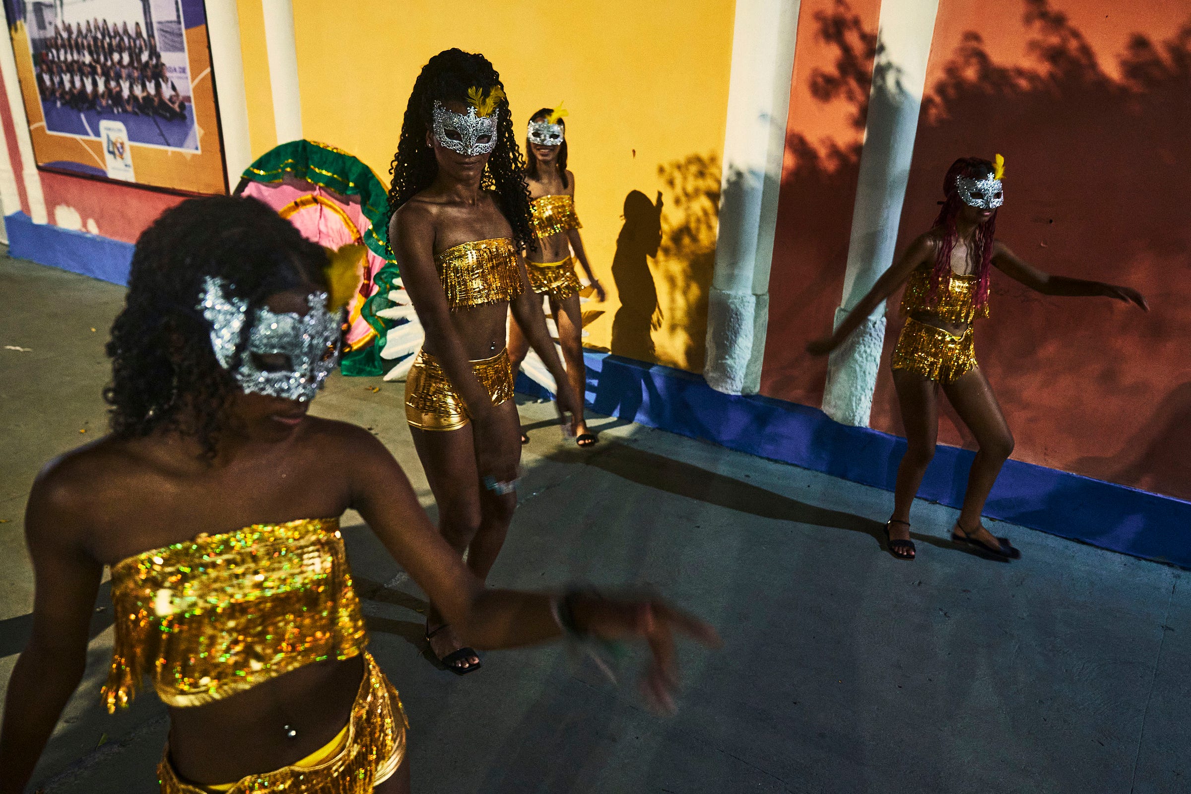 Four people in gold sequin outfits and silver masks dance outdoors against a yellow and orange wall, casting long shadows on the pavement in the evening light.