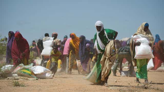 A woman dragging a large sack as other women around her walk away with white sacks picked up from the dusty ground. A woman dragging a large sack as other women around her walk away with white sacks picked up from the dusty ground.