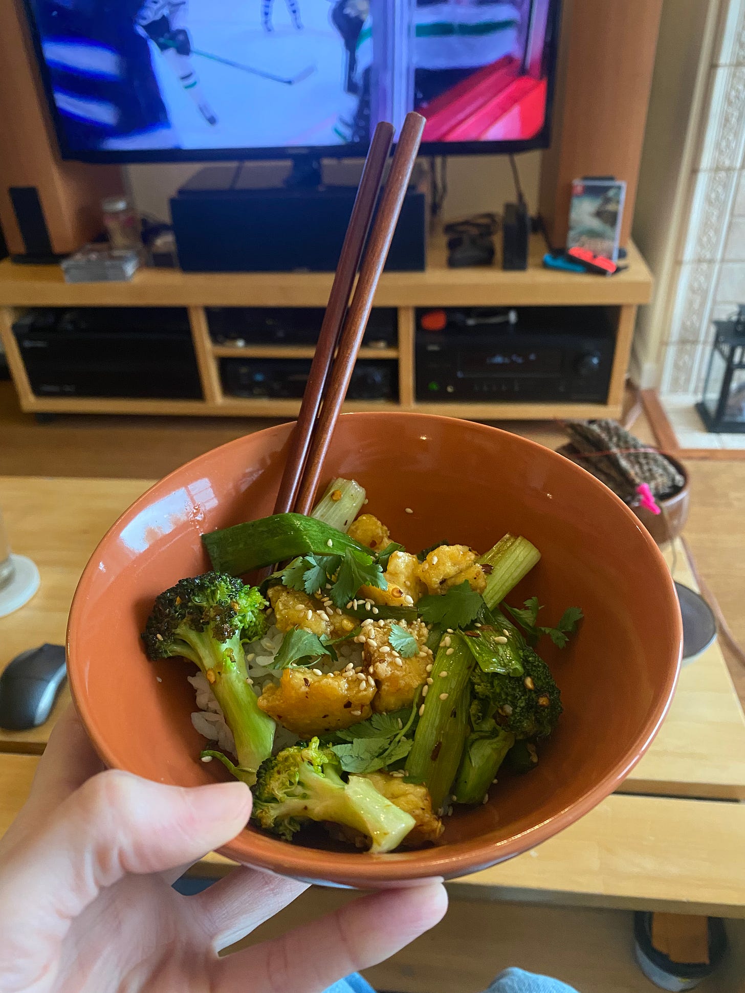 My hand hold an orange bowl of rice and the tofu/broccoli/green onion mix described above, topped with sesame seeds and cilantro. In the background is the coffee table, and the tv showing a hockey game.