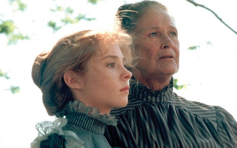 Anne, her hair in a loose bun, stands next to Marilla, hair also in a bun. Both wear high-necked, gray-striped dresses and gaze seriously into the distance.