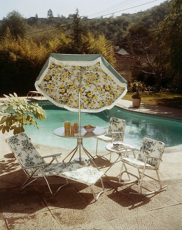 Floral patio furniture around a pool with a matching umbrella. Drinks and chips on a table, set in a sunlit backyard with lush trees.