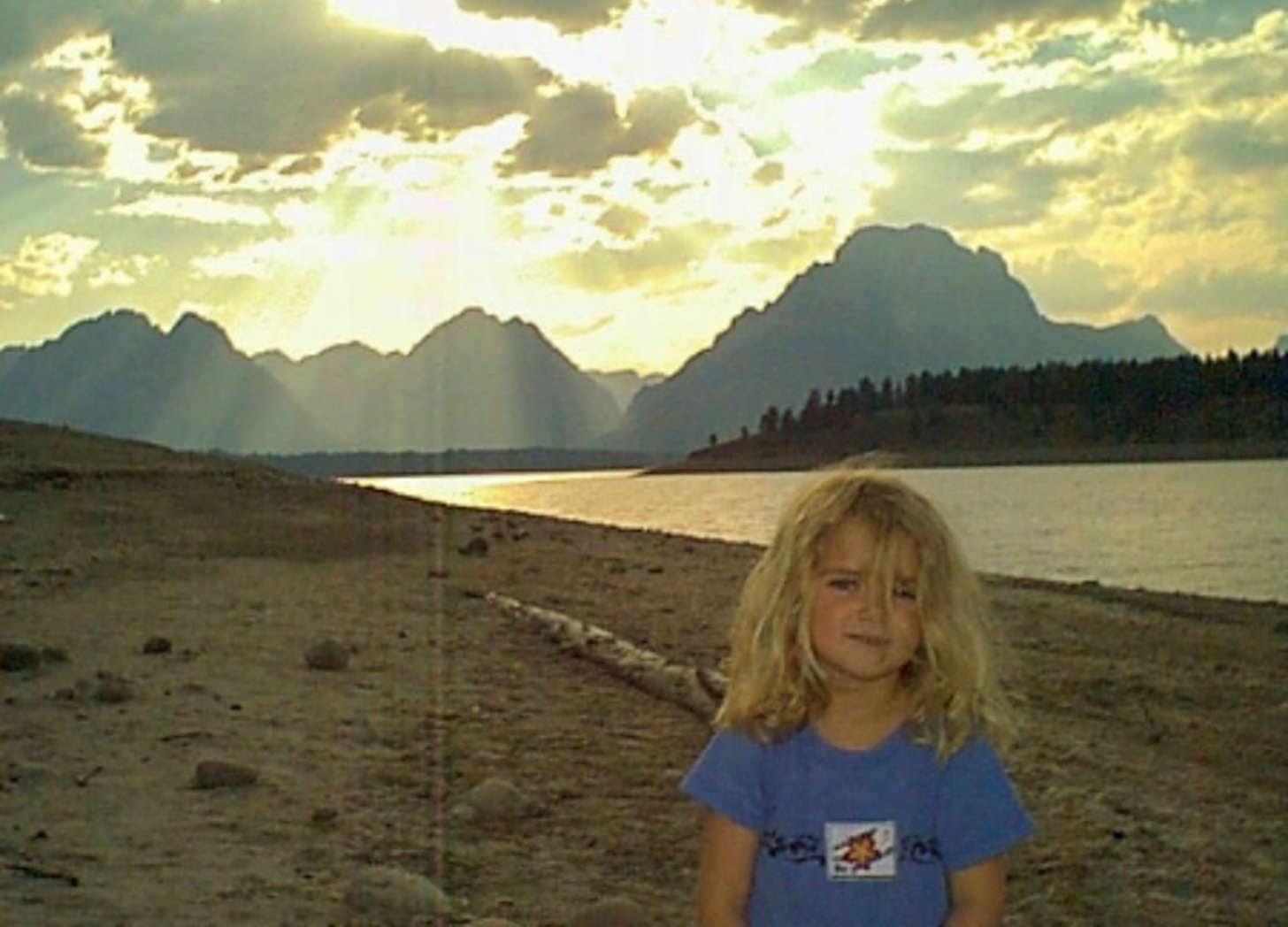 Little girl at shore of lake with Grand Tetons in background at sunset