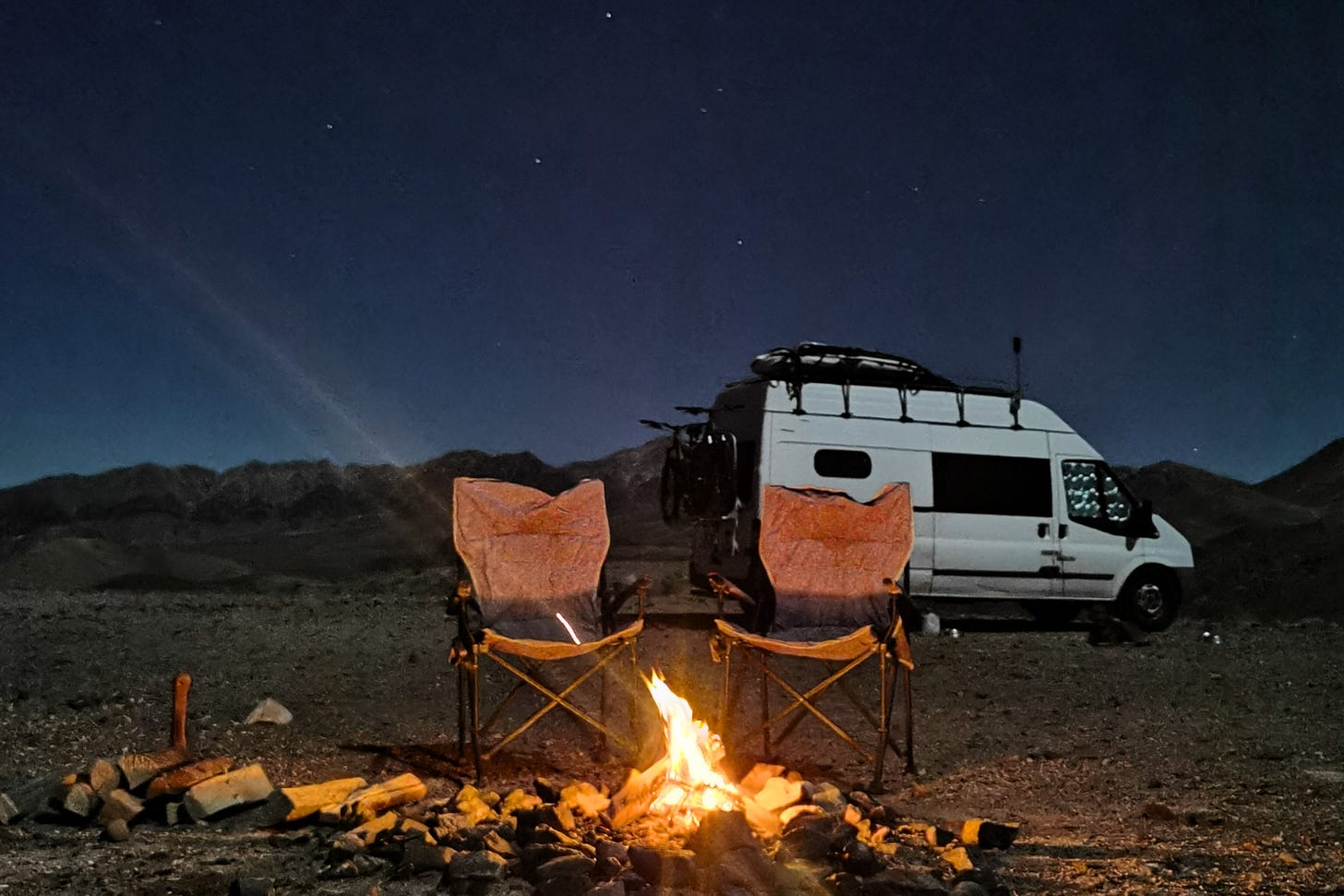 Two empty camp chairs sit in front of a glowing campfire under a starry sky, with a white camper van parked nearby. The scene is set in a remote, mountainous desert landscape, capturing the quiet solitude and warmth of vanlife after dark. Two empty camp chairs sit in front of a glowing campfire under a starry sky, with a white camper van parked nearby. The scene is set in a remote, mountainous desert landscape, capturing the quiet solitude and warmth of vanlife after dark.
