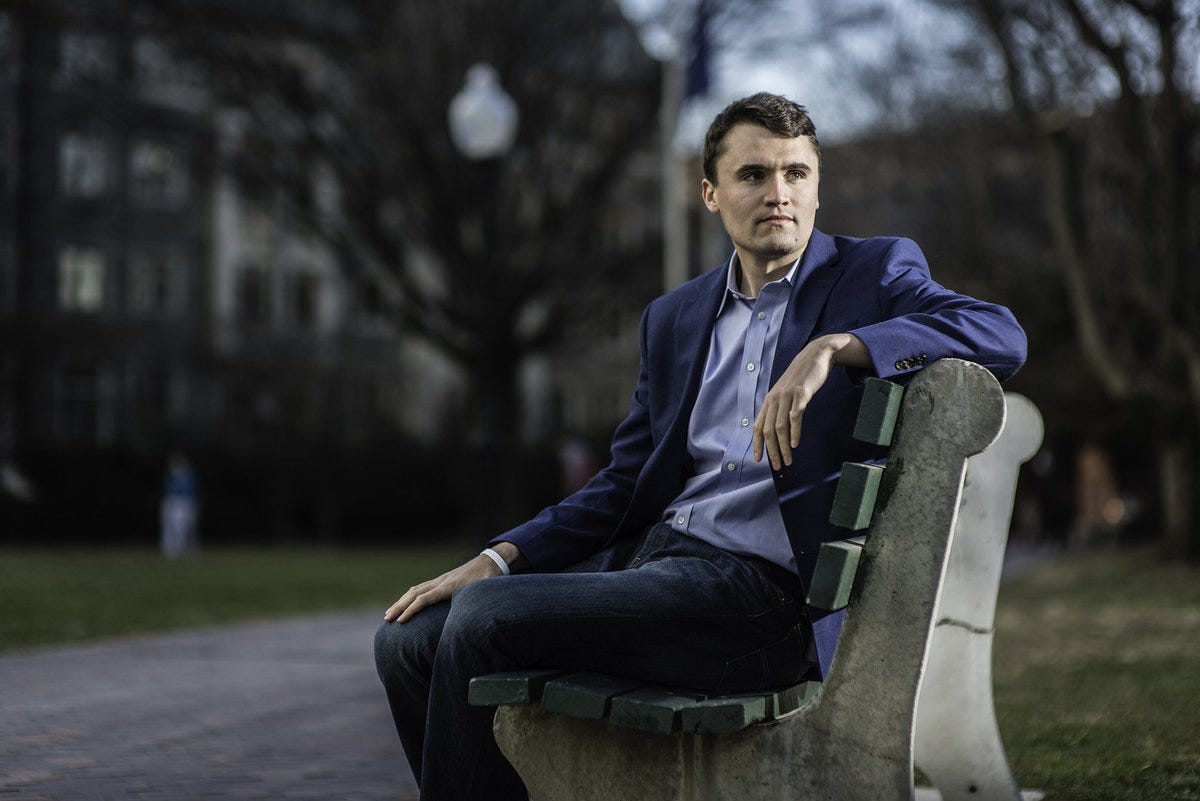 Eli Steele sitting on a bench in a park. He wears a blue suit jacket, light blue shirt, and dark jeans. Trees and a pathway are visible in the background.