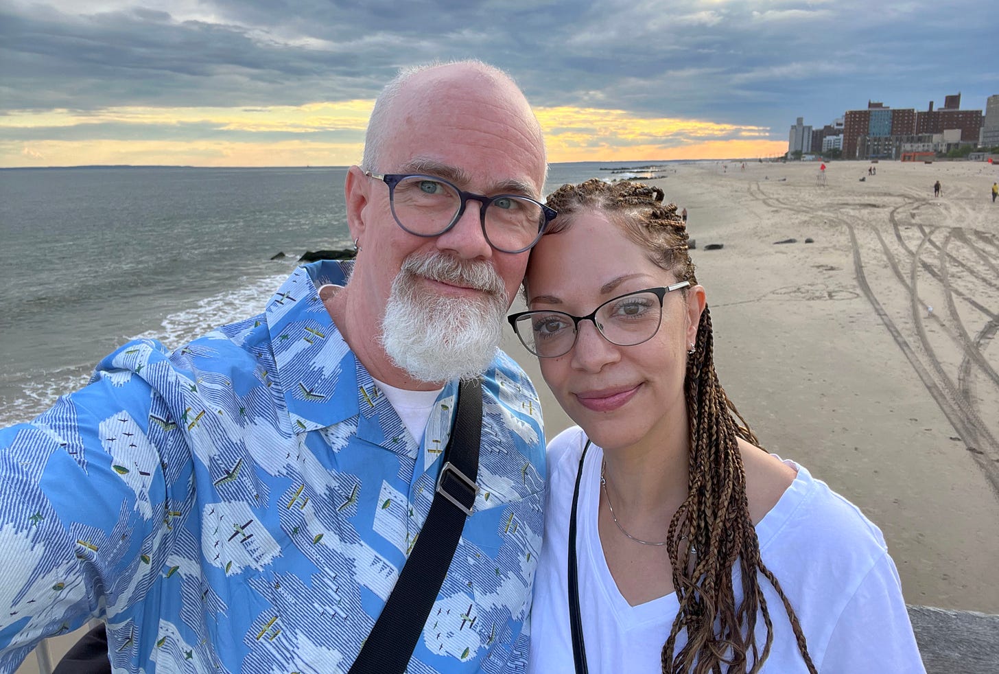 A man and woman lean together taking a selfie on a cloudy beach near dusk. He is bald with a gray goatee. She has long braids pulled back and hanging over her shoulder.